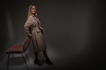 young, pretty, emotional girl in a gray raincoat standing near a chair, leaning on a white background, a ray of light