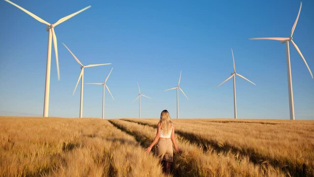 Young blonde woman walks through a golden wheat field with wind turbines on the horizon and dreams of a clean and sustainable future for humanity