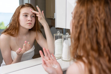Red-haired girl applies cream to her face looking at herself in the bathroom mirror