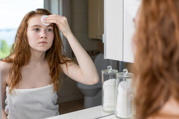 Red-haired teenage girl wipes her face with lotion after shower looking at herself in the mirror