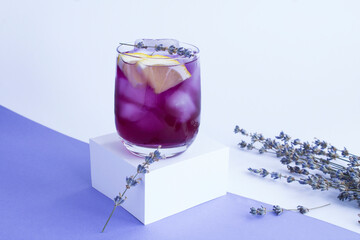Lavender lemonade with ice and lemon in the drinking glass on the white cube on the colored...