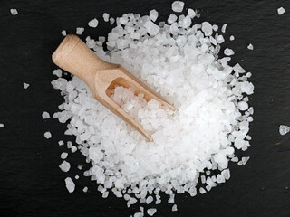 wooden spice scoop on heap of coarse sea salt on black slate stone, top view of natural clear salt...
