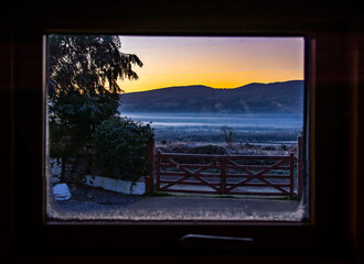 View through the window of a colourful sunrise at the countryside with frosty fence and misty background