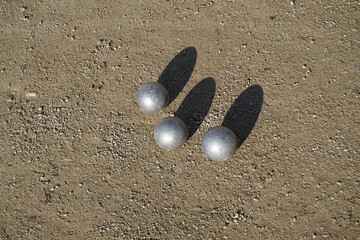 Drei silberne Boulekugeln auf braunem feuchten Erdboden bei Sonne am Mittag im Winter
