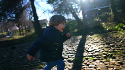 One happy small boy running outside in city park wearing jacket and boots. Excited child runs outdoors in sunlight