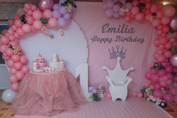 Festive decorations, pink balloons arch, white chair in crown shape, wooden number one. Birthday cake and candy bar. 1 year old girl birthday party photo zone