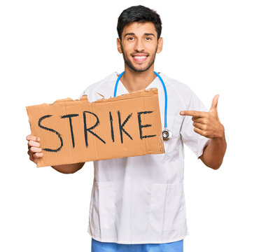 Young handsome man wearing doctor uniform holding strike banner cardboard smiling happy pointing with hand and finger