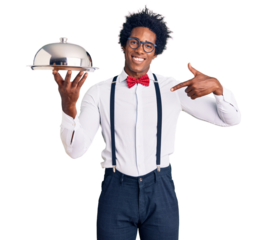 Handsome african american man with afro hair wearing waiter uniform holding silver tray pointing finger to one self smiling happy and proud