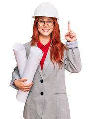 Young redhead architect woman wearing safety helmet holding blueprints surprised with an idea or question pointing finger with happy face, number one