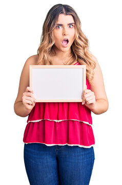 Young caucasian woman holding empty white chalkboard scared and amazed with open mouth for surprise, disbelief face