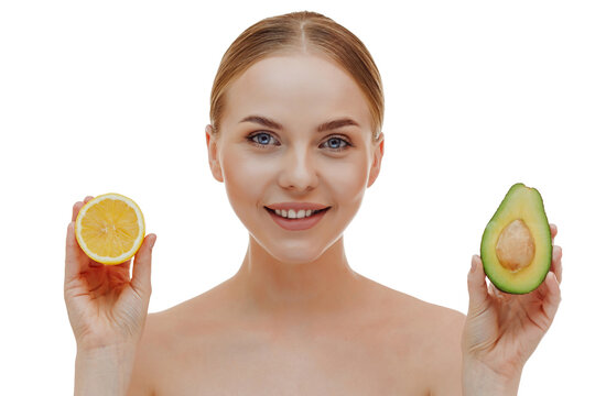 Close-up shot of a pretty young woman gently touching her face, having a well-groomed complexion, healthy glowing skin, standing with bare shoulders, holding half a juicy lemon and half an avocad