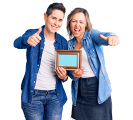 Couple of women holding empty frame approving doing positive gesture with hand, thumbs up smiling and happy for success. winner gesture.