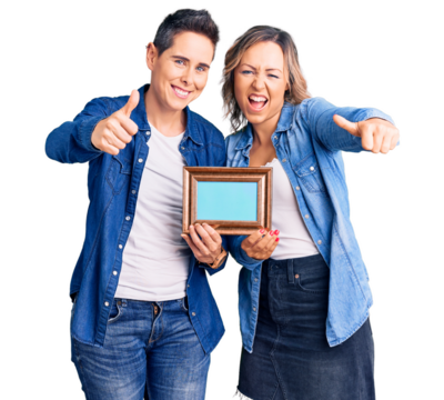 Couple of women holding empty frame approving doing positive gesture with hand, thumbs up smiling and happy for success. winner gesture.