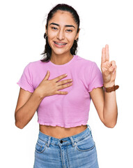 Hispanic teenager girl with dental braces wearing casual clothes smiling swearing with hand on...