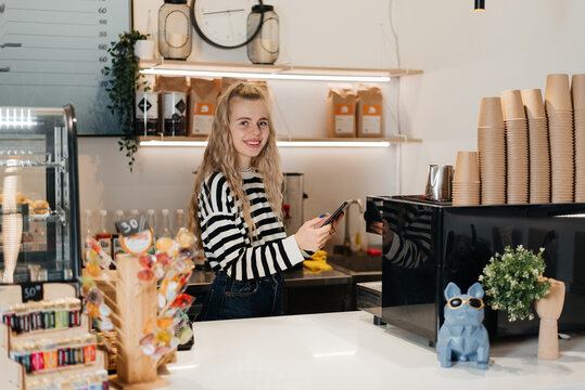 Beautiful Young Caucasian Barista with Blond Hair is Making a Cup of Fresh Coffee in a Cafe. Happy and Smiling Bar Employee Posing while Working in a Cozy Loft-Style Coffee Shop Restaurant Counter. - Powered by Adobe