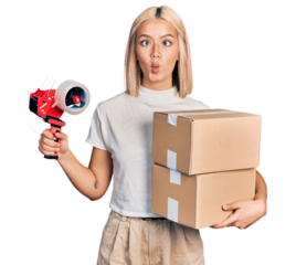 Beautiful young blonde woman holding packing tape and cardboard boxes making fish face with mouth and squinting eyes, crazy and comical.