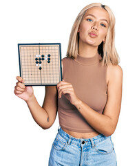 Beautiful young blonde woman holding asian go game board looking at the camera blowing a kiss being...