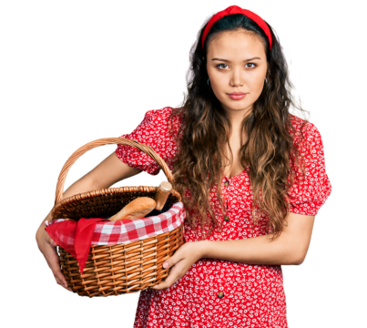 Young hispanic girl holding picnic wicker basket with bread relaxed with serious expression on face. simple and natural looking at the camera.