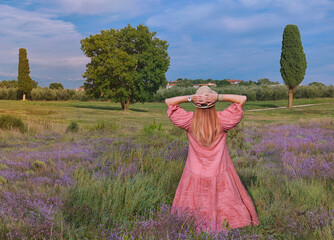 Back view of happy woman with hands up in straw hat  on a lavender field. Landscape with a cypress tree near the Green lagoon sea bay in Porec, Croatia - Istria, Europe - 576440850