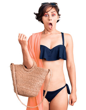 Beautiful young woman with short hair wearing bikini and hat holding summer wicker handbag scared and amazed with open mouth for surprise, disbelief face