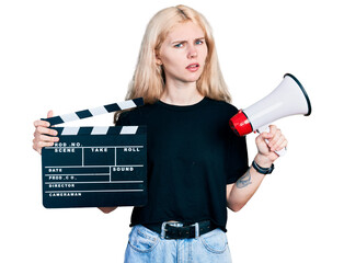 Young caucasian woman holding video film clapboard and megaphone clueless and confused expression....