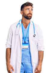 Young hispanic man wearing doctor uniform and stethoscope smiling looking to the side and staring away thinking.