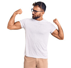 Young hispanic man wearing casual clothes and glasses showing arms muscles smiling proud. fitness...