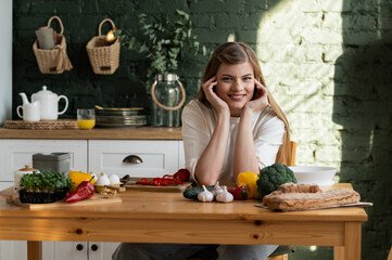 Photo on the topic of healthy eating. A young woman in the kitchen sits at a wooden table and looks to the side. Various vegetables, products and greens with dishes are on the table.