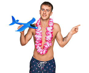 Young blond man wearing swimsuit and hawaiian lei holding airplane toy smiling happy pointing with...