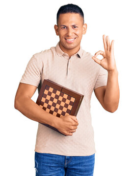 Young handsome latin man holding chess doing ok sign with fingers, smiling friendly gesturing excellent symbol