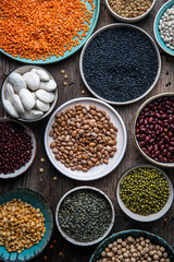 Different legumes (lentils, beans, chickpeas, mung, peas) in bowls and plates on wooden background.