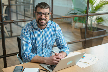 Portrait of smiling man sitting at his desk in the office, using laptop
