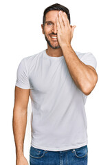 Handsome man with beard wearing casual white t shirt covering one eye with hand, confident smile on face and surprise emotion.