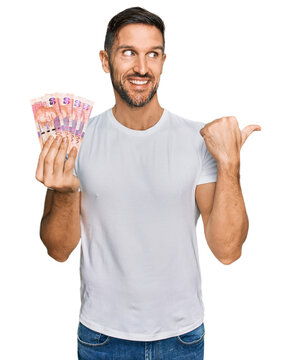 Handsome man with beard holding south african 50 rand banknotes pointing thumb up to the side smiling happy with open mouth