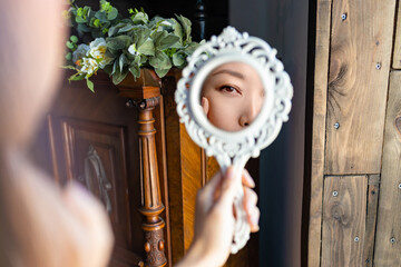 beautiful young asian woman looks at herself in the mirror, hand on cheek.