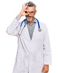 Middle age grey-haired man wearing doctor uniform and stethoscope smiling happy doing ok sign with hand on eye looking through fingers