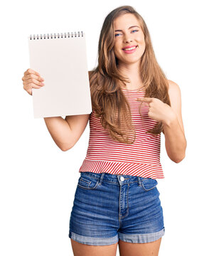 Beautiful caucasian young woman holding notebook pointing finger to one self smiling happy and proud