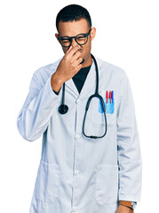 Young african american man wearing doctor uniform and stethoscope smelling something stinky and disgusting, intolerable smell, holding breath with fingers on nose. bad smell