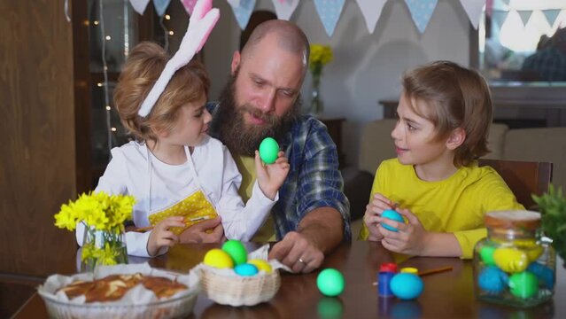 Easter Family traditions. Father and two caucasian happy children with bunny ears dye and decorate eggs with paints for holidays while sitting togeKids embrace and smile in cozy.