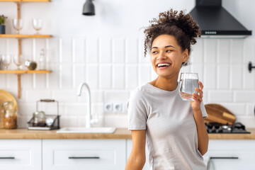 Portrait of a beautiful African American young woman drinking water standing in the kitchen at home looking away and smiling, wellness healthy food concept - Powered by Adobe