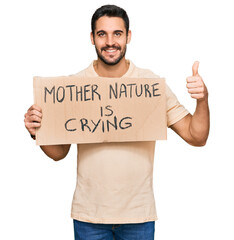 Young hispanic man holding mother nature is crying protest cardboard banner smiling happy and positive, thumb up doing excellent and approval sign