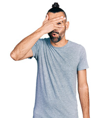 Hispanic man with ponytail wearing casual grey t shirt peeking in shock covering face and eyes with hand, looking through fingers with embarrassed expression.