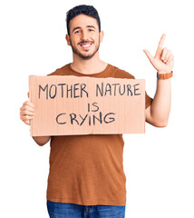 Young hispanic man holding mother nature is crying protest cardboard banner surprised with an idea or question pointing finger with happy face, number one