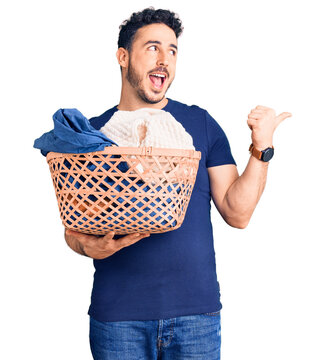 Young hispanic man holding laundry basket pointing thumb up to the side smiling happy with open mouth