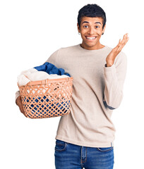 Young african amercian man holding laundry basket celebrating victory with happy smile and winner...