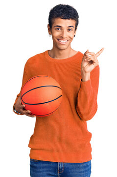 Young african amercian man holding basketball ball smiling happy pointing with hand and finger to the side