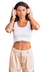 Young hispanic woman with tattoo listening to music using headphones thinking attitude and sober expression looking self confident