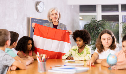 Mature female teacher working in a high school tells pupils the history of Austria in lesson and holds the national flag of the .country in her hands