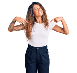 Young hispanic woman with tattoo wearing casual white tshirt looking confident with smile on face,...