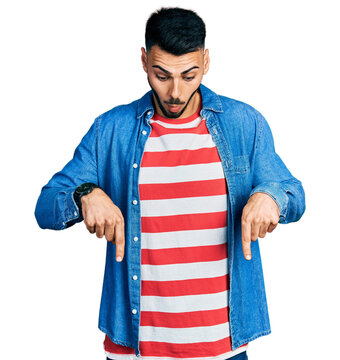 Young hispanic man with beard wearing casual denim jacket pointing down with fingers showing advertisement, surprised face and open mouth